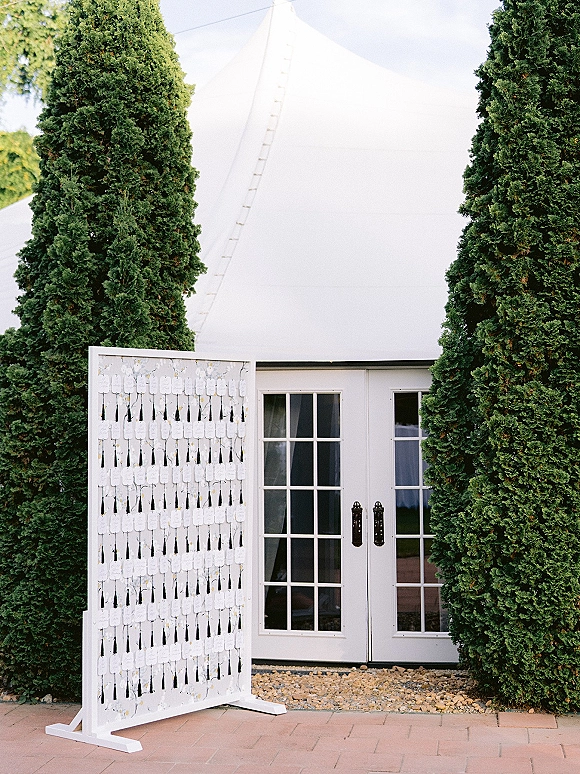 Wedding escort display with escort card wall of hanging calligraphy tags and tassels on a wooden frame easel by a white tent entrance