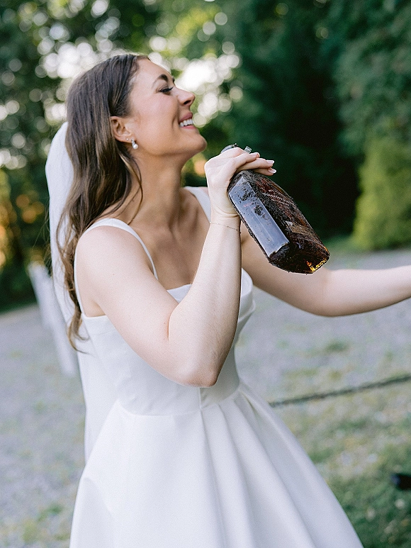 Bride portrait of a laughing bride drinking from a bottle, wearing a veil and satin wedding dress on a gravel path by trees