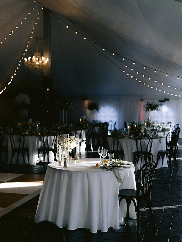 Reception tablescape with round wedding tables in white linens, taper candles and white florals under draped tent ceiling with string lights
