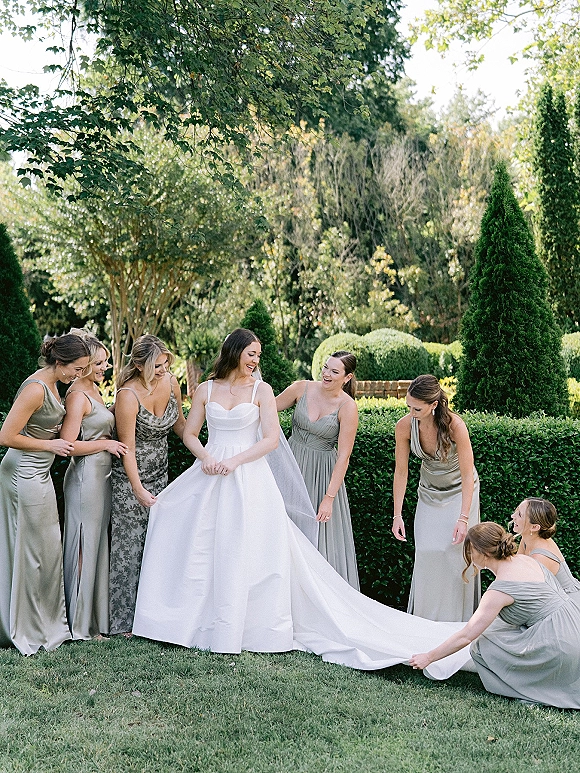 Bride with bridesmaids in a bridal party portrait as they kneel to fix her long dress train, veil accent, amid garden hedges and trees