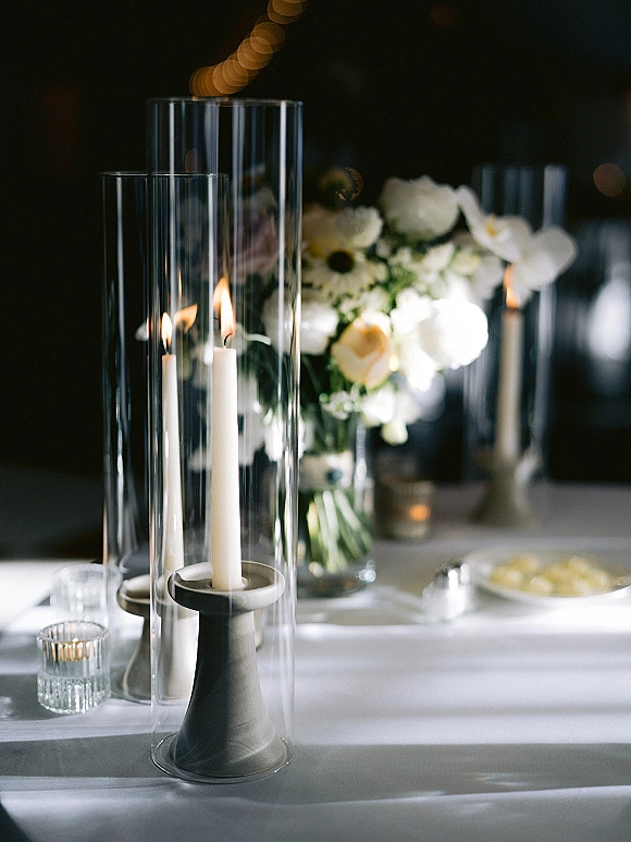 Reception tablescape with wedding table candles, white taper and glass hurricane candles around white flowers and greenery under string lights
