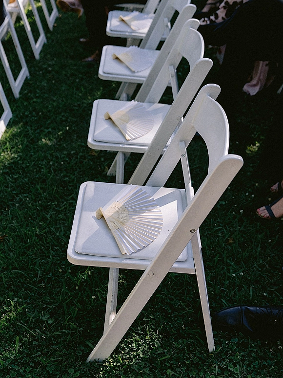 Ceremony seating with outdoor ceremony chairs, white folding chairs lined on a grass lawn with paper hand fans on each seat
