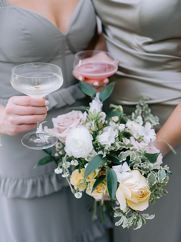 Bridesmaid toast with bridesmaids holding drinks, raising champagne coupe glasses beside rose and greenery bouquets against a neutral backdrop