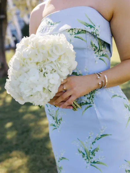 Bridal bouquet of white hydrangeas held against a strapless floral dress, with ring and bracelets visible in sunlit lawn setting