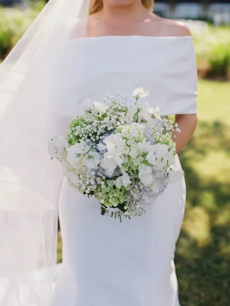 Bridal bouquet of white hydrangeas with baby’s breath and green foliage held by a bride in an off-the-shoulder gown on a sunny lawn
