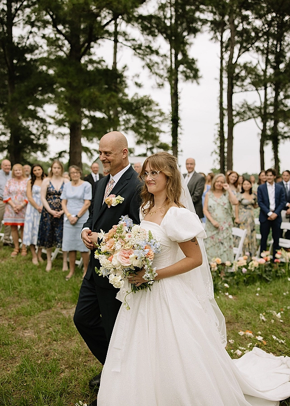 Processional moment as bride walking down aisle in veil and glasses, holding bouquet, escorted past petals and guests on a tree-lined lawn