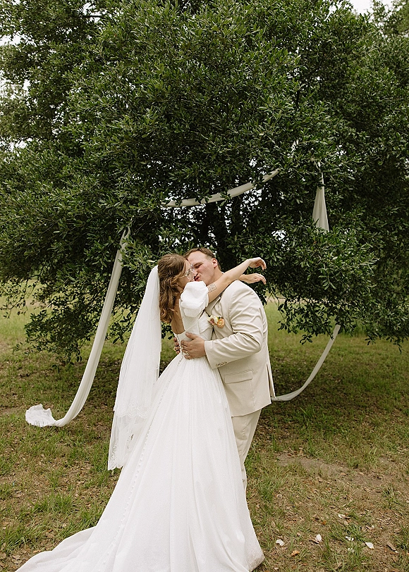 Wedding kiss portrait of bride and groom kissing beneath a fabric-draped arch, her long veil flowing on a grassy lawn under trees