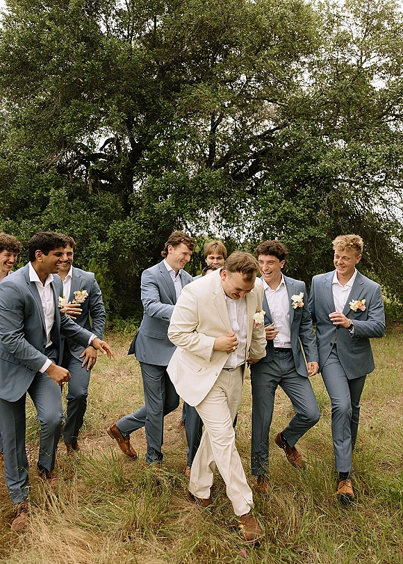 Groomsmen portrait of the groom with groomsmen in light blue suits and a cream suit, walking and laughing in a grassy field by trees