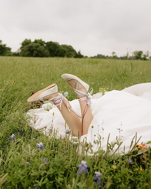 Bridal shoes, white wedding flats with pastel ribbon laces resting on wedding dress fabric in tall grass of a wildflower meadow