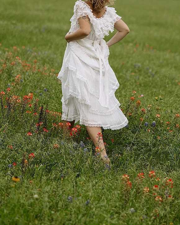 Bridal portrait of a bride in wildflowers wearing a lace wedding dress with ruffle sleeves and layered skirt, standing in a meadow