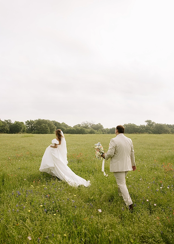 Couple portrait of bride and groom from behind walking through a wildflower meadow, her veil and dress train trailing, groom holding bouquet
