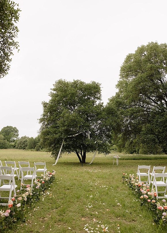 Outdoor ceremony setup with a garden ceremony aisle of white folding chairs and flower petals, framed by rose bushes in a grassy field
