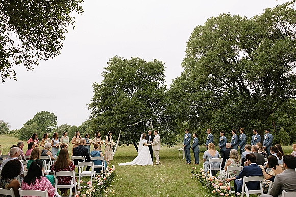 Wedding ceremony under an oak tree with fabric-draped arch, flower-lined aisle, and guests seated on white chairs in a meadow field