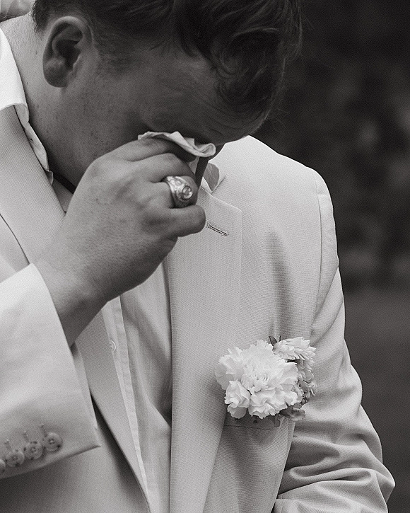 Emotional groom portrait of a groom wiping tears, wearing a light suit with boutonniere, against soft outdoor greenery backdrop