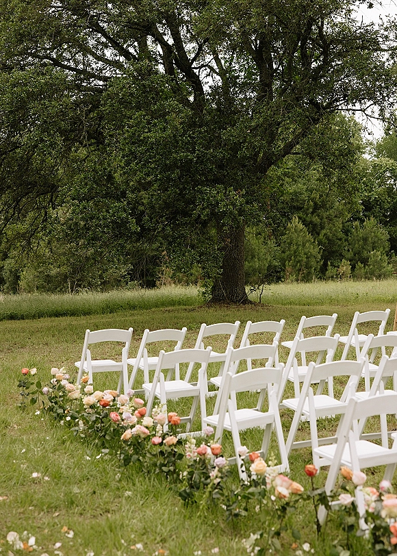 Ceremony setup with white folding chairs in rows and rose aisle arrangements on a grass lawn beneath a large tree in a garden setting