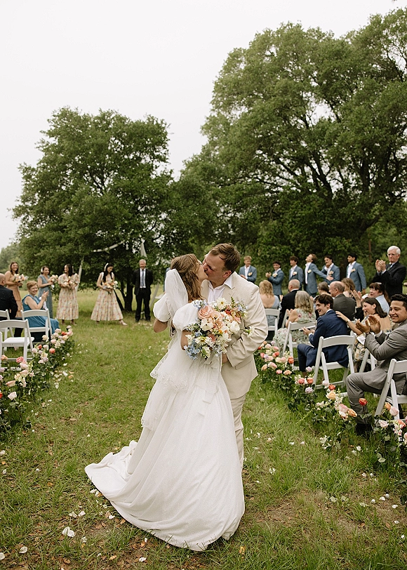 Ceremony kiss at an outdoor wedding ceremony as the bride and groom kiss in the aisle, bouquet and veil amid rose petals and cheering guests