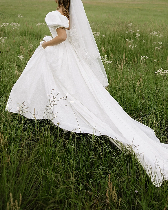 Bridal portrait of a bride in field with a long wedding dress train and lace veil flowing over meadow grass and wildflowers