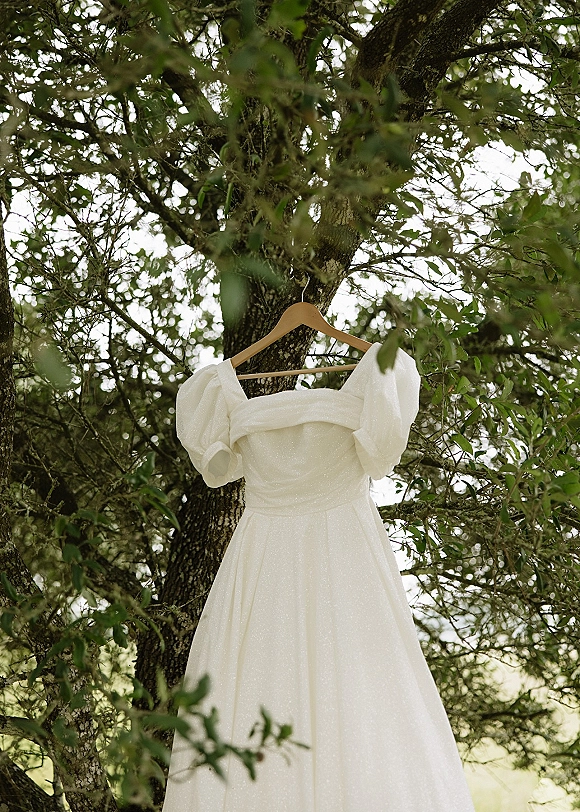 Wedding dress hanging from a wooden hanger, off the shoulder neckline and puff sleeves in satin, framed by green garden tree branches