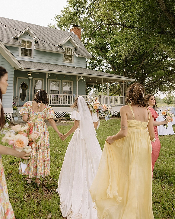 Bridal party walking on a lawn, bride in veil and gown with wildflower bouquet alongside bridesmaids in mismatched dresses by a farmhouse porch