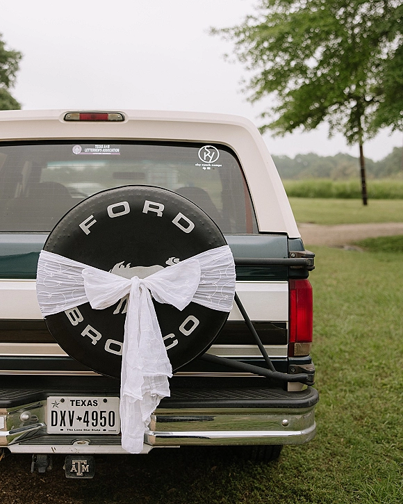 Wedding getaway car with a white tulle bow on the rear spare tire, parked on a gravel drive by a grassy lawn under cloudy sky