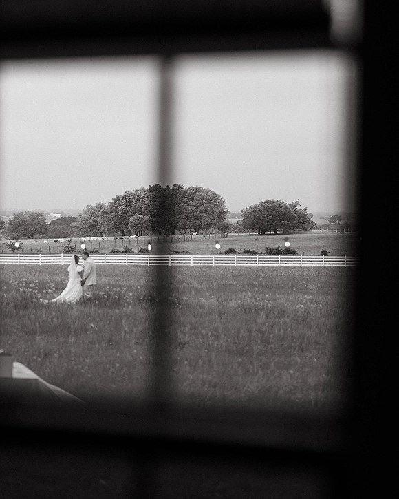 Wedding couple portrait of bride and groom in field, framed by a window, her veil and dress train flowing by a white fence and trees