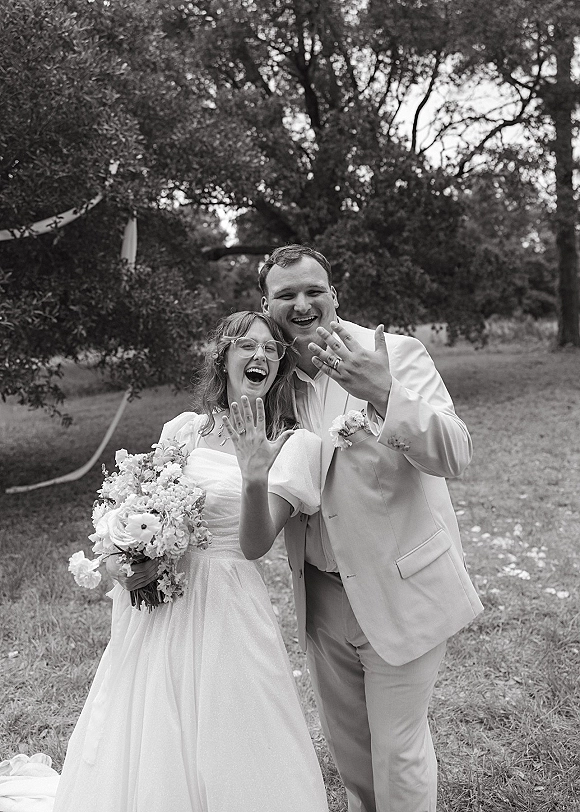 Couple portrait in a black and white wedding photo, bride and groom laughing while showing rings, bouquet in hand on a garden path