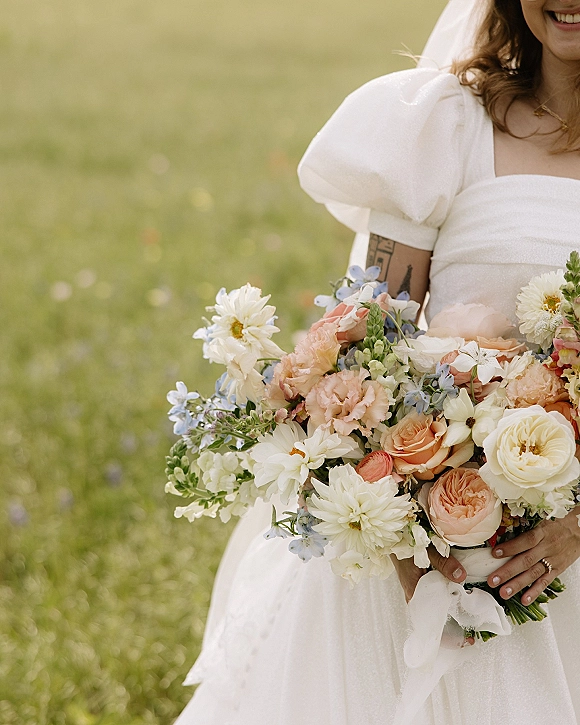 Bridal bouquet of pastel wildflowers in peach and cream, ribbon-wrapped in the bride’s hands with ring detail, in a green meadow