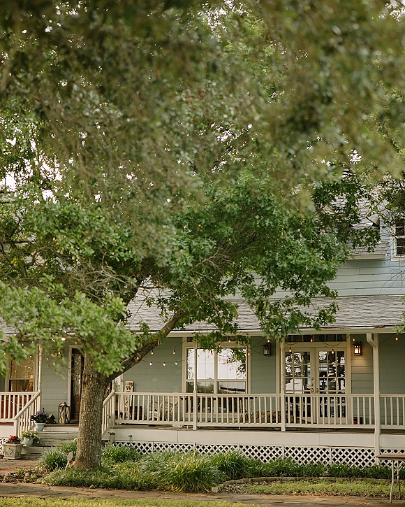 Wedding venue exterior with a farmhouse wedding venue porch, string lights and lanterns glowing beside flower pots under a large tree canopy
