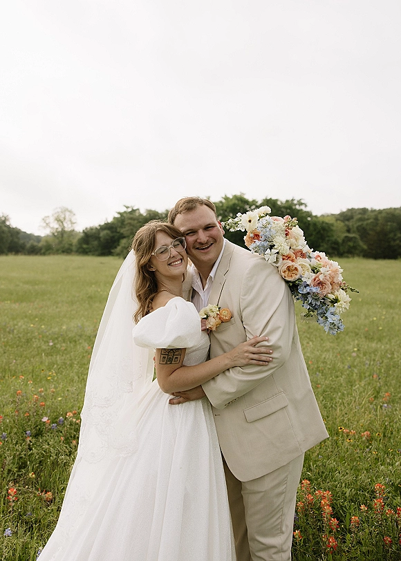 Couple portrait of bride and groom hugging with a blue and peach bouquet, veil and glasses in a wildflower meadow under overcast sky