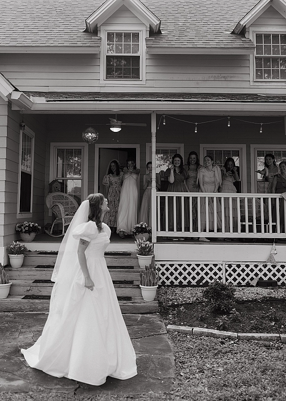 Bride reveal moment as bridesmaids reaction unfolds on a house porch, bride in veil and full skirt gown under string lights with disco ball decor