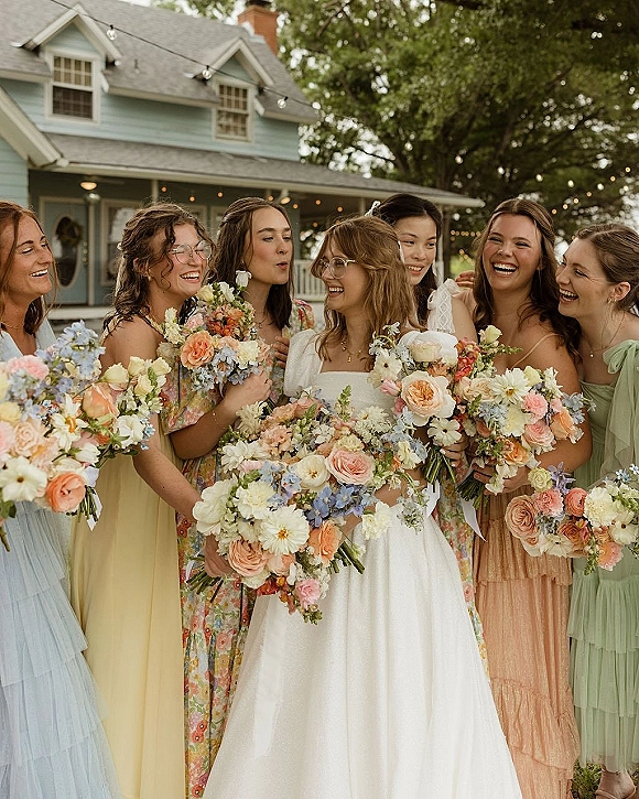 Bridesmaid group photo with bride and bridesmaids holding pastel wedding bouquets on a farmhouse porch, one in eyeglasses under string lights