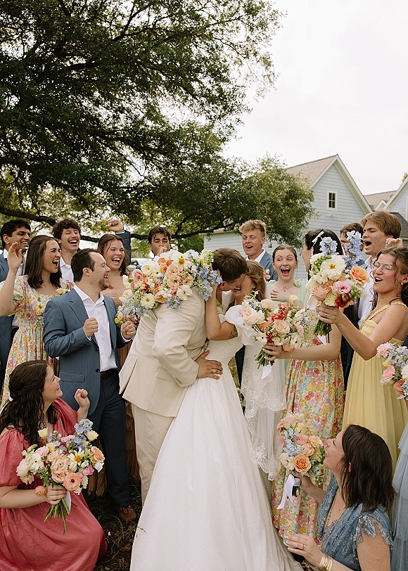 Wedding party photo of the bride and groom kissing as the bridal party cheers, holding bouquets under tree shade near a farmhouse lawn