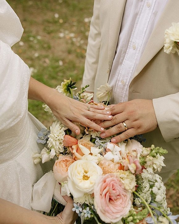 Wedding ring exchange as the bride slides a gold band onto the groom’s finger, bouquet ribbon and boutonniere accents over outdoor grass backdrop