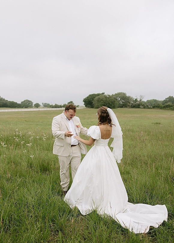 First look moment as bride in a cathedral veil walks up to groom in an ivory suit, holding hands in a meadow field with fence under cloudy sky