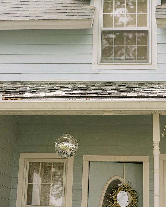 Front porch decor with a hanging disco ball and wreath on a light blue front door under a covered porch beside house windows