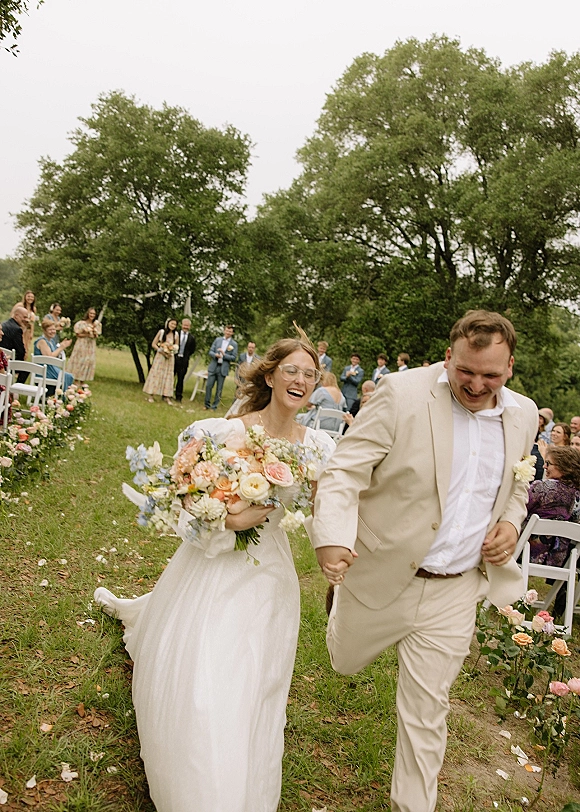 Recessional moment as bride and groom running hand in hand down the aisle, bride with bouquet and glasses, guests cheering in a grassy field