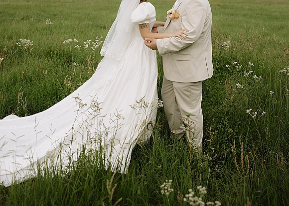 Couple portrait of bride and groom embrace in a wildflower meadow, her long veil and train flowing as they hold a peach rose bouquet