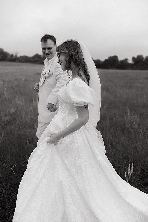 Couple portrait of bride and groom walking hand in hand, bride holding her dress with veil flowing through a meadow field under open sky
