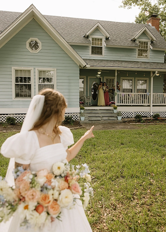 Bride portrait holding bouquet, pointing as bridesmaids on a farmhouse porch hold bouquets behind her under string lights