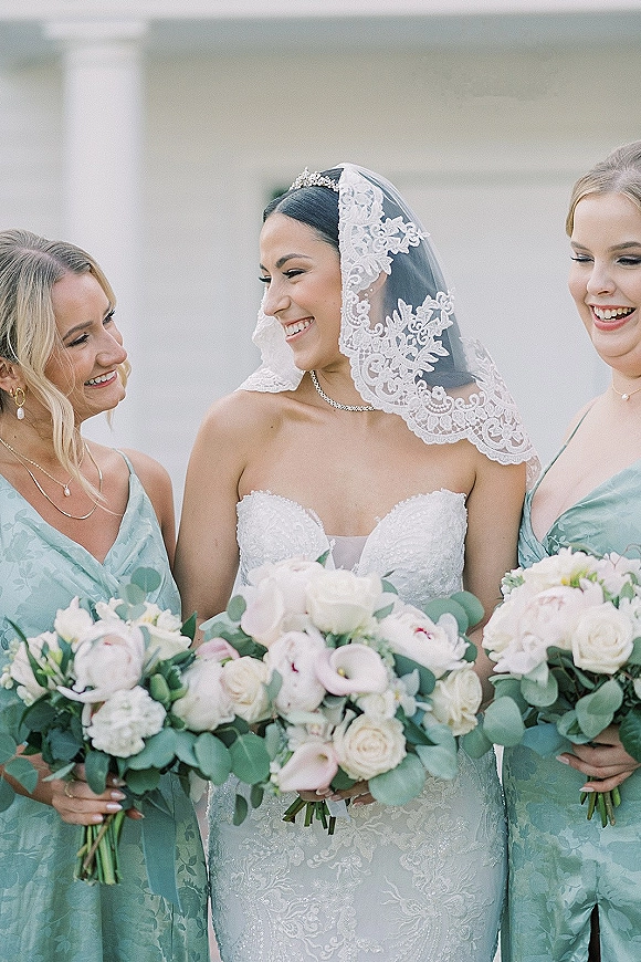 Bride with bridesmaids portrait featuring a laughing bride in lace veil and tiara, holding white and blush bouquets by a light exterior wall