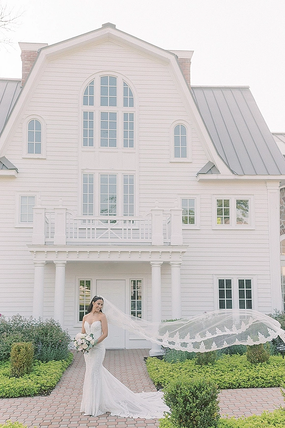 Bridal portrait of a bride with long veil holding a bouquet, veil trailing along a brick walkway outside a white house with shrubs