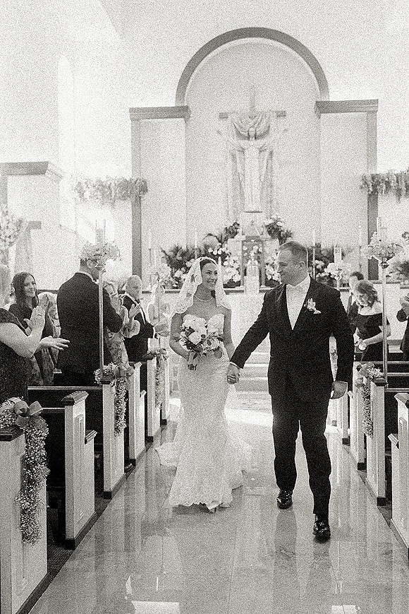 Wedding recessional as bride and groom walk hand in hand down a white aisle runner in a church, bride with bouquet and veil, guests behind