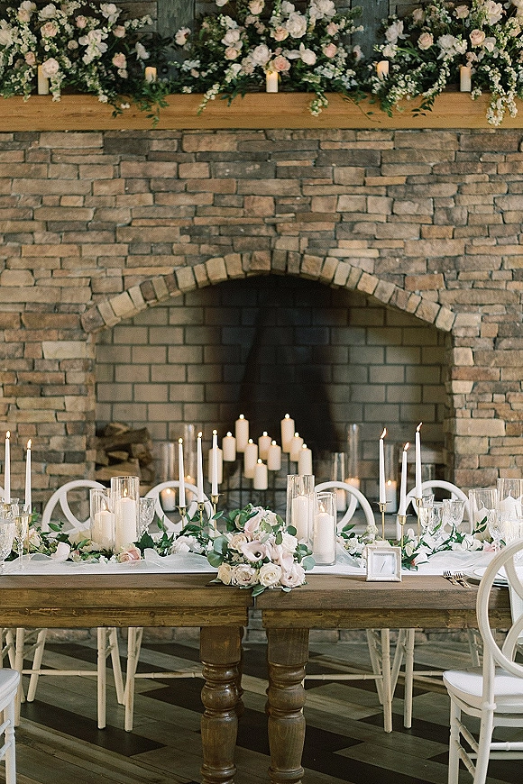 Reception tablescape with blush roses and greenery garland, taper candles in gold holders and hurricanes, set before a stone fireplace