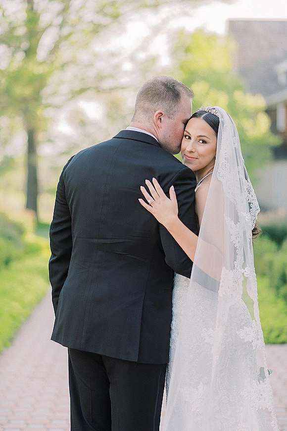Couple portrait of groom kissing bride’s forehead as she looks over her shoulder, lace veil accent, on a tree-lined outdoor walkway near a building