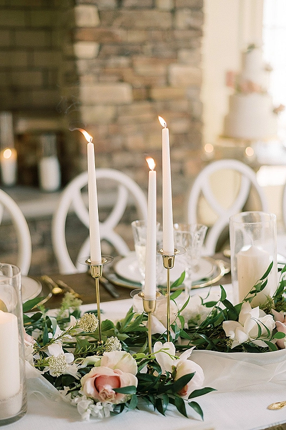 Reception tablescape with wedding table centerpiece of taper candles in brass candlesticks and greenery garland, set before a stone fireplace