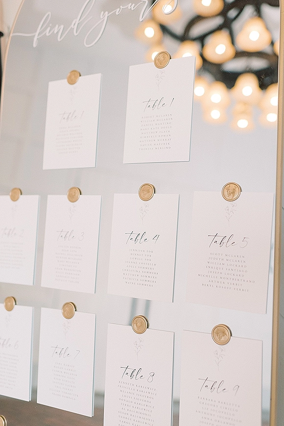 Wedding seating chart mirror with gold frame and hanging seating cards sealed with wax, glowing softly against string light bokeh