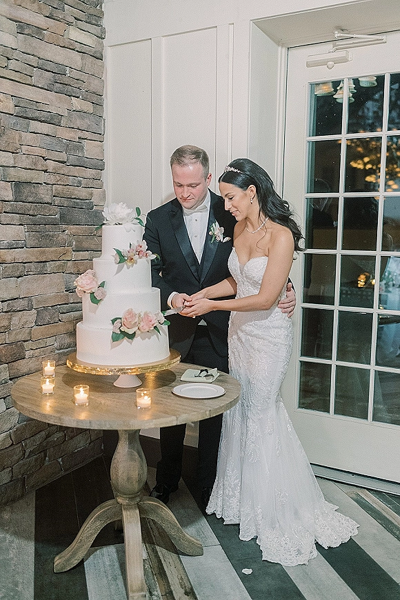 Wedding cake cutting as bride and groom cut a three-tier buttercream cake with sugar flowers at a candlelit table by a stone wall