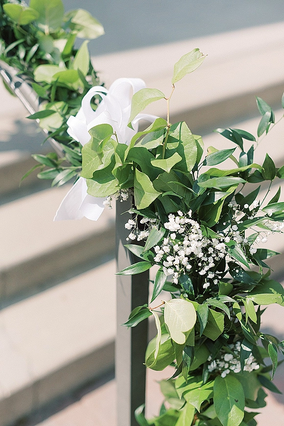 Aisle floral decor with wedding aisle greenery and baby's breath garland tied with white ribbon along a metal railing on sunlit stone steps