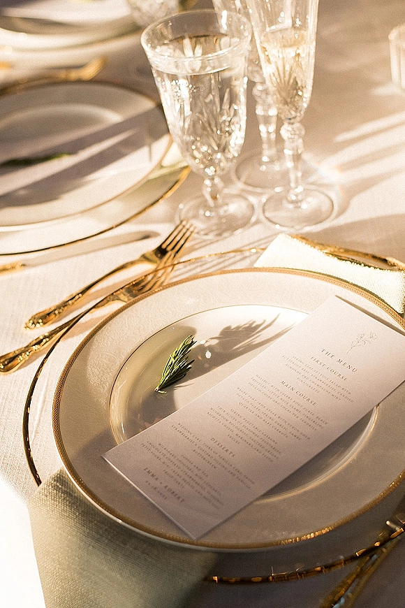 Reception tablescape with wedding place setting of gold rim plates, crystal champagne flutes, menu card and rosemary sprig in sunlit shadows