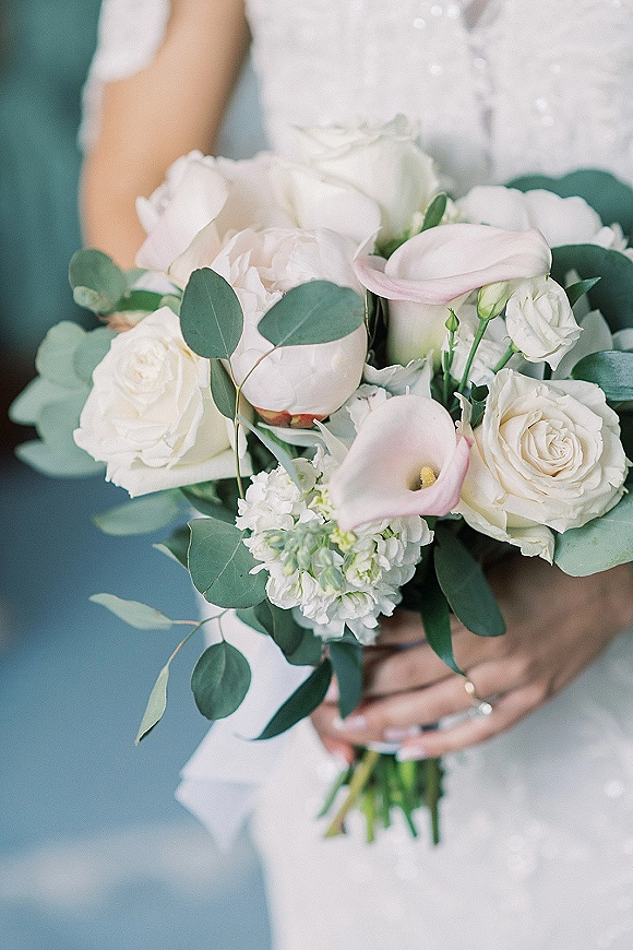 Bridal bouquet of white roses and peonies with eucalyptus greenery, held by a bride in a gown showing her engagement ring against a neutral backdrop